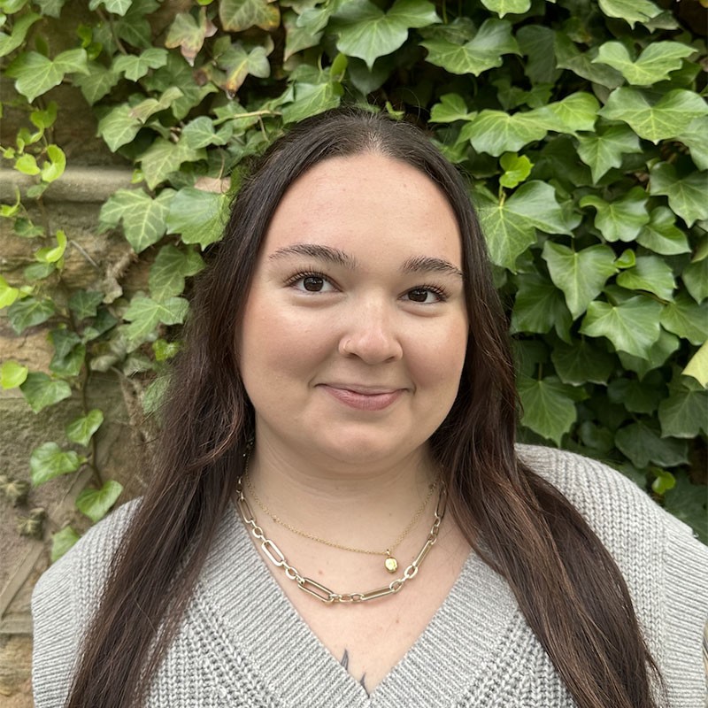 Portrait of Baylie McMahon, standing outdoors in front of a lush green ivy-covered wall. Baylie is wearing a gray knit sweater and a gold chain necklace, and has long, dark hair.